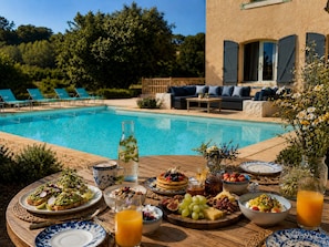 A poolside breakfast setup with a variety of food items, a glass of orange juice, and a view of the pool and outdoor seating area.