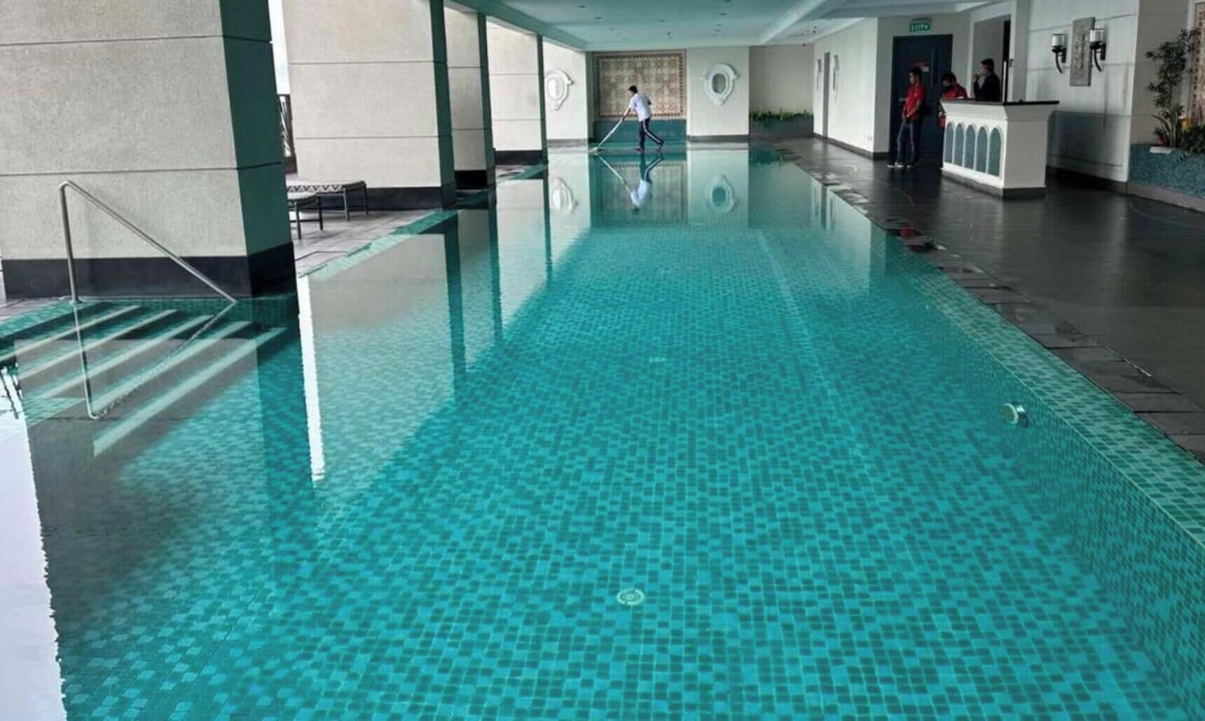 A large indoor swimming pool with clear blue water, a reception desk, and a person cleaning the pool.