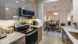 A modern kitchen with stainless steel appliances, a dining area with a round table, and a hallway leading to a living space. - The Breeze at Lewes Condo w/ Pool | Bike to Beach! (Lewes)