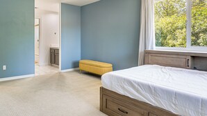 Un dormitorio con cama, ventana con cortinas, un banco amarillo y una puerta que da a otra habitación.