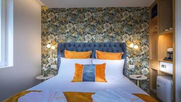 Camera d'albergo con un letto grande, carta da parati floreale, due lampade ai lati del letto e un tavolino con un frigorifero.