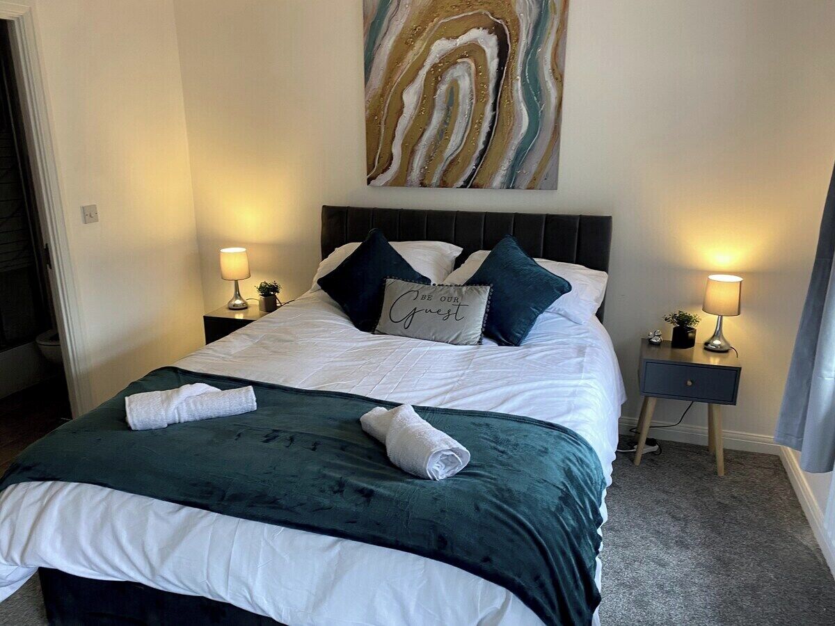 Un letto rifatto con cura, un cuscino con la scritta 'BE OUR GUEST' e una coperta verde. Due comodini con lampade e un'opera d'arte appesa sopra il letto.