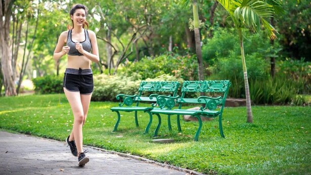 Una persona trotando por un sendero pavimentado en un parque con bancos verdes y árboles.