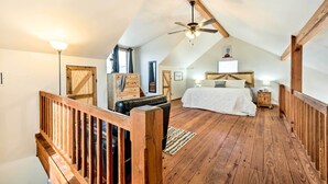 Quarto com teto inclinado, vigas de madeira, cama, mesa de cabeceira e ventoinha.