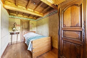 Ein hölzernes Bett in einem Raum mit Holzboden, ein kleiner Tisch mit einer Lampe und eine Holztür.