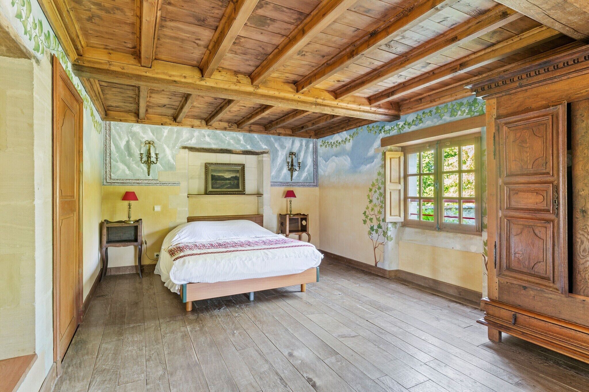 A bedroom with a wooden ceiling, a bed with a white and red bedspread, two bedside tables with lamps, a large wooden wardrobe, and a mural on the wall.