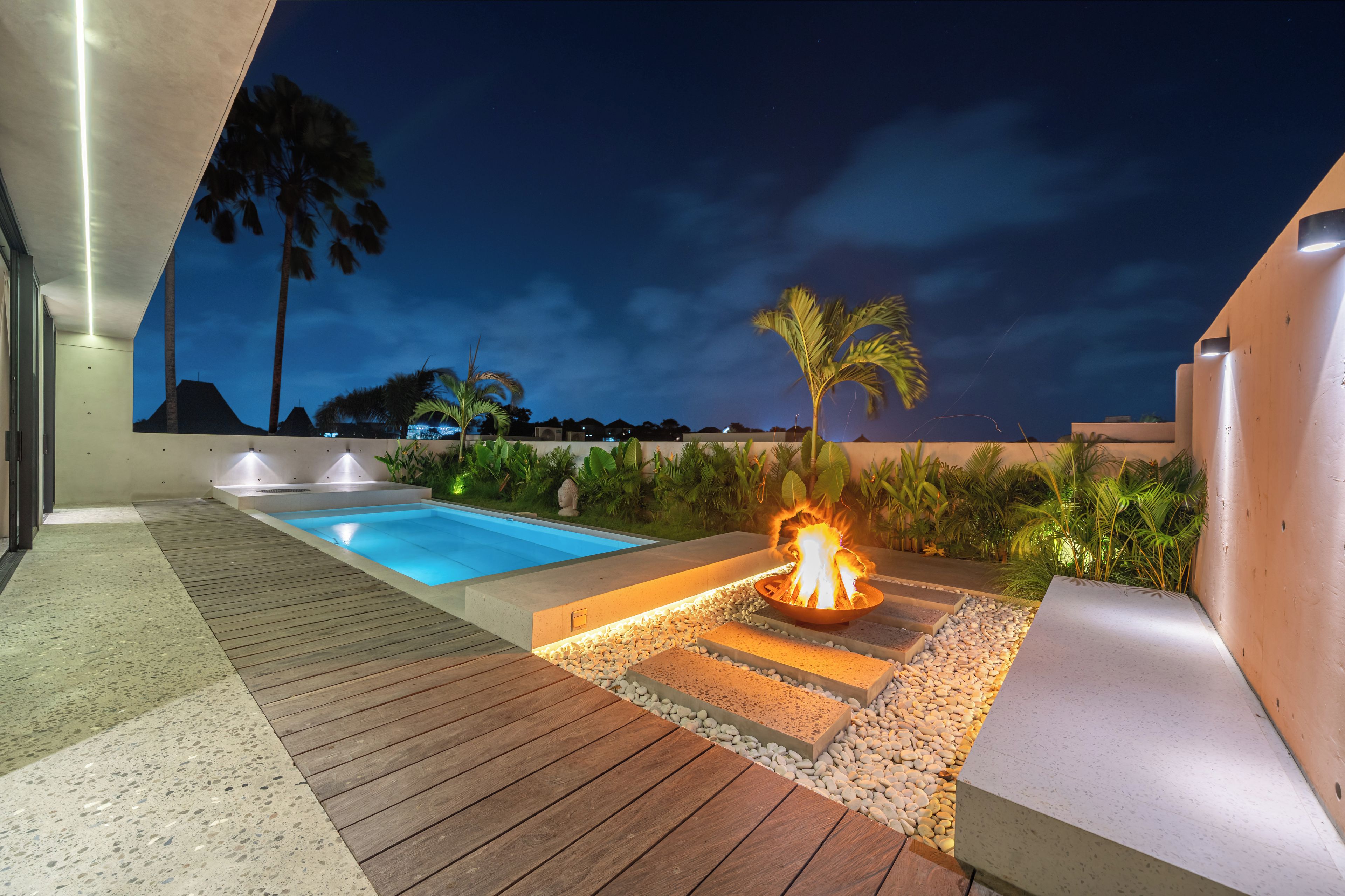 Zona de piscina exterior de noche con una chimenea, cubierta de madera y vegetación circundante.