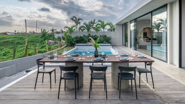 Un área de comedor al aire libre con piscina, rodeada de plantas tropicales y un edificio moderno.