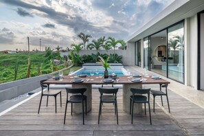 Ett utomhusområde med plats för måltider, en pool och tropiska växter, omgivet av en modern byggnad.