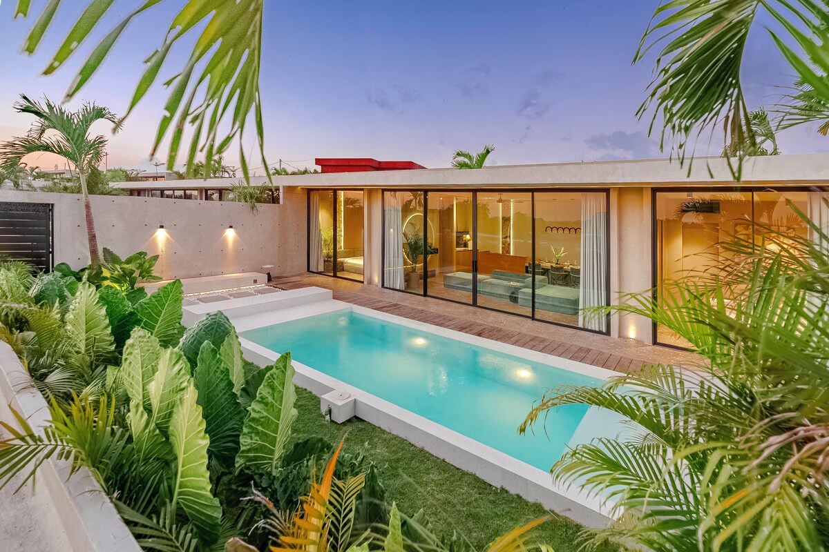 Un área moderna junto a la piscina con una sala con paredes de vidrio, abundante vegetación y cielo despejado.
