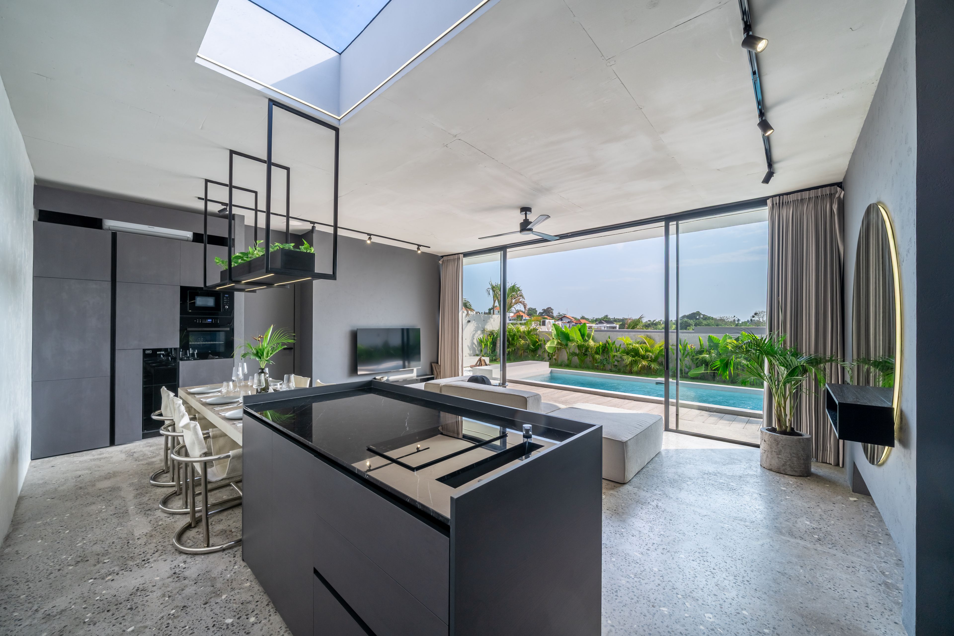 Une cuisine moderne dotée d’un îlot central intégré, d’un coin repas avec une table à plateau en verre et d’une grande porte-fenêtre coulissante donnant sur une piscine.