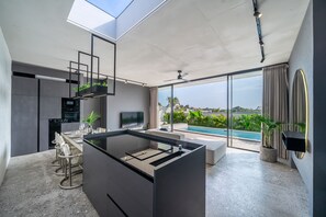 A modern kitchen with a built-in island, a dining area with a glass top table, and a large sliding glass door leading to a pool. - Predmet Cascade Bali (Tabanan)