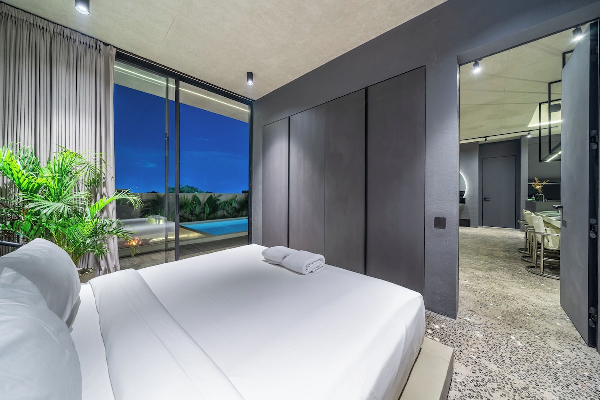 Una habitación de hotel moderna con una cama grande, vista a una piscina por la noche y una zona de comedor separada.