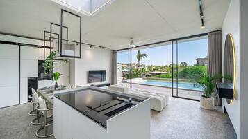 A modern kitchen with a central island, a dining area with a pool view, and a living space with a large window overlooking the outdoors.