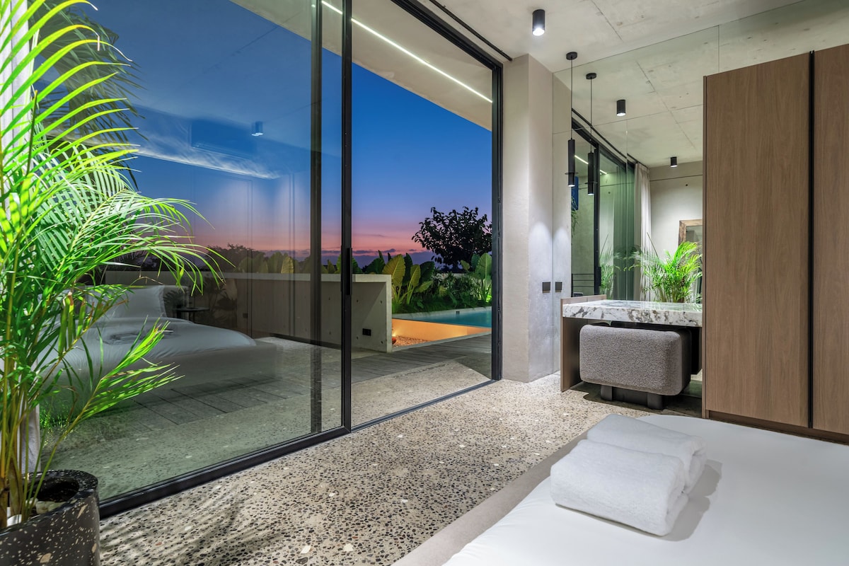 Un baño moderno con un amplio ventanal de vidrio que da a un balcón, una encimera de mármol y una planta en maceta.