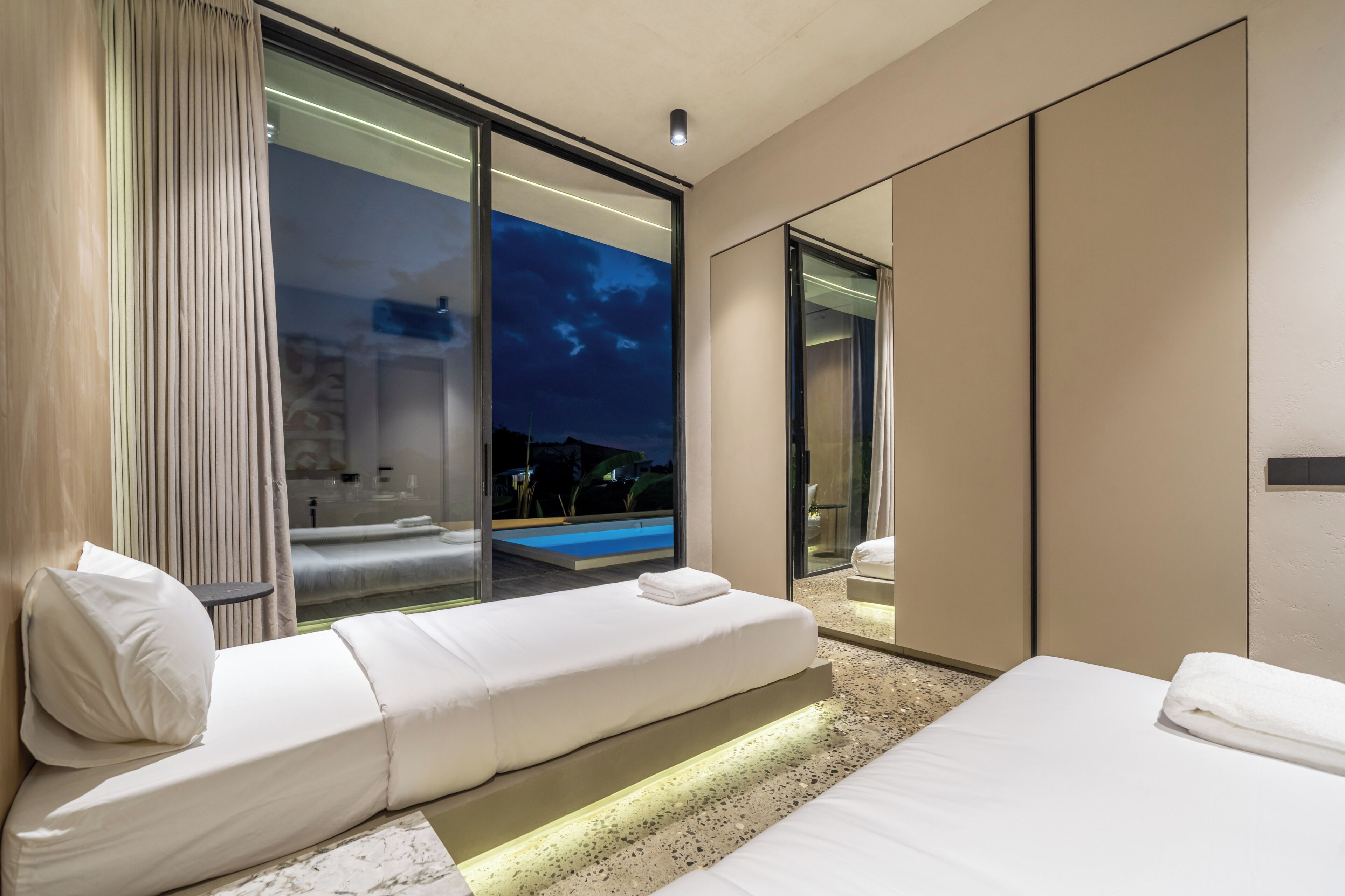 A modern hotel room with a large bed, a seating area with a view of an outdoor pool, and a glass wall.