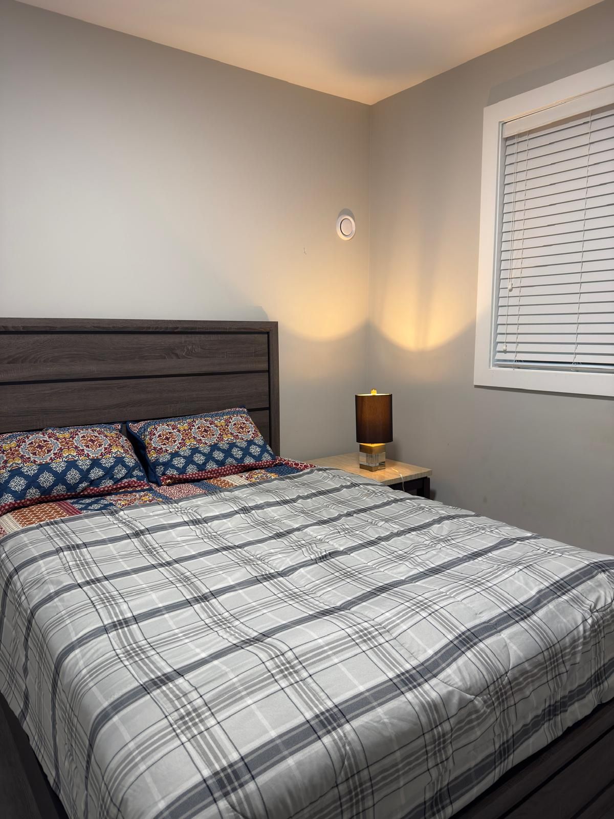 Un dormitorio con cama, mesita de noche, lámpara, luz empotrada en la pared y ventana con persianas.