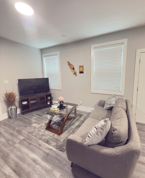 A living room with a flat-screen TV, a glass coffee table, a grey sofa, and a decorative wall piece. - PORTSMOUTH COVE! Peaceful North Nanaimo Retreat. (Nanaimo)