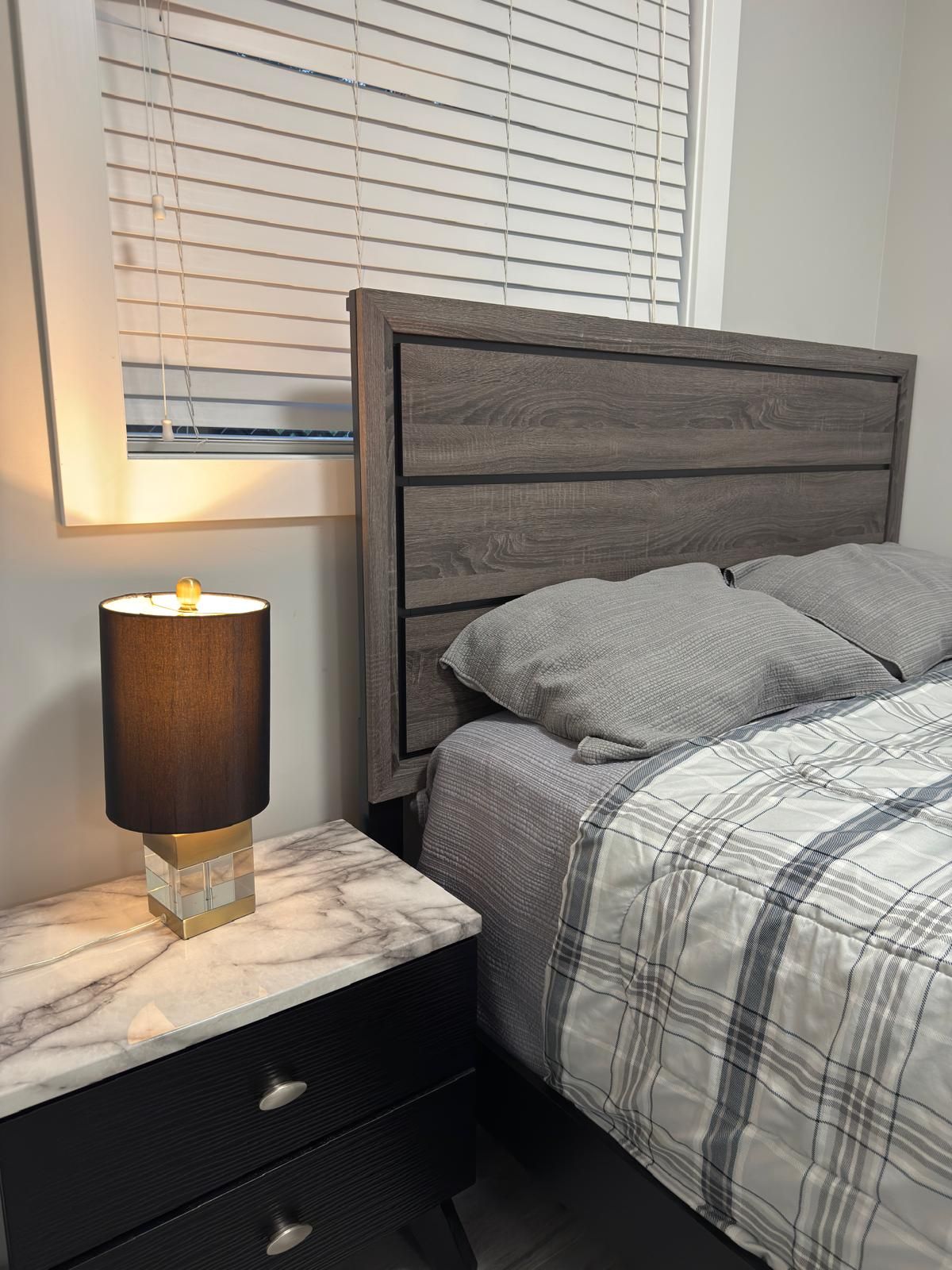 A bedroom with a bed, a nightstand with a lamp, and a window with blinds.