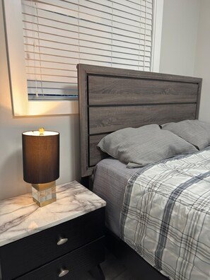 Et soveværelse med en seng, et natbord med en lampe og et vindue med rullegardiner.