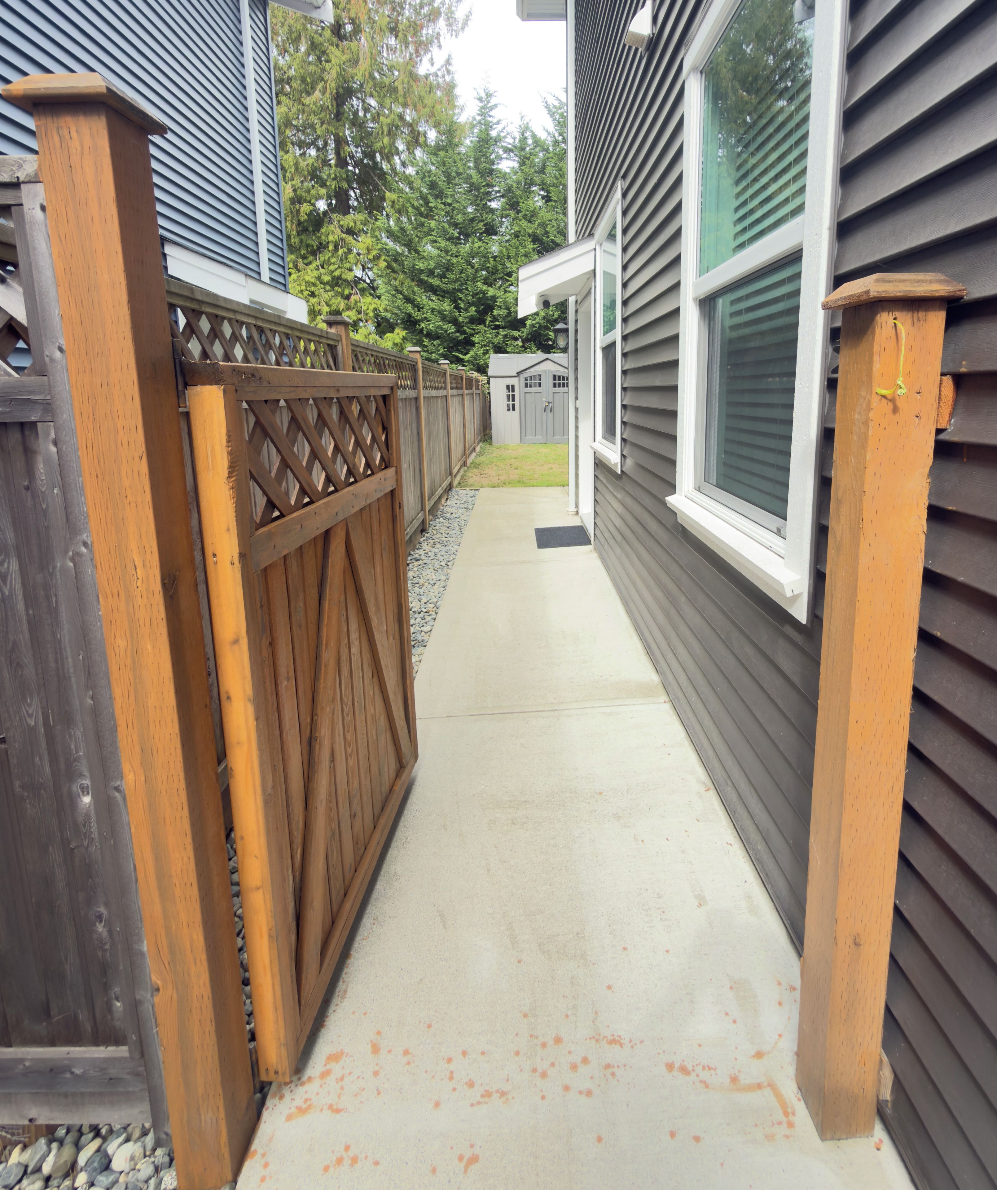 Un chemin de béton mène à un portail en bois, de part et d’autre duquel se trouve une maison avec un revêtement extérieur et une fenêtre.