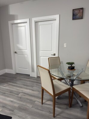 Een eetruimte met een glazen tafel, vier stoelen, twee witte deuren en een kleine potplant aan de muur.