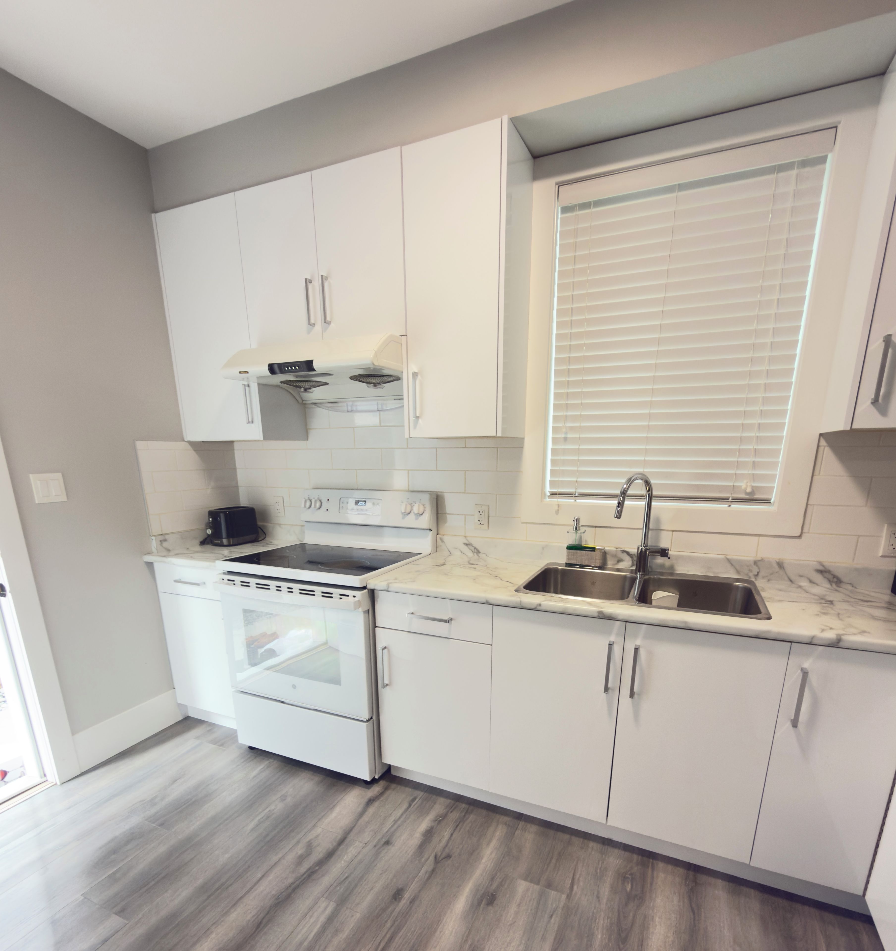 A kitchen with white cabinets, a stove, an oven, a sink, and a window with blinds.