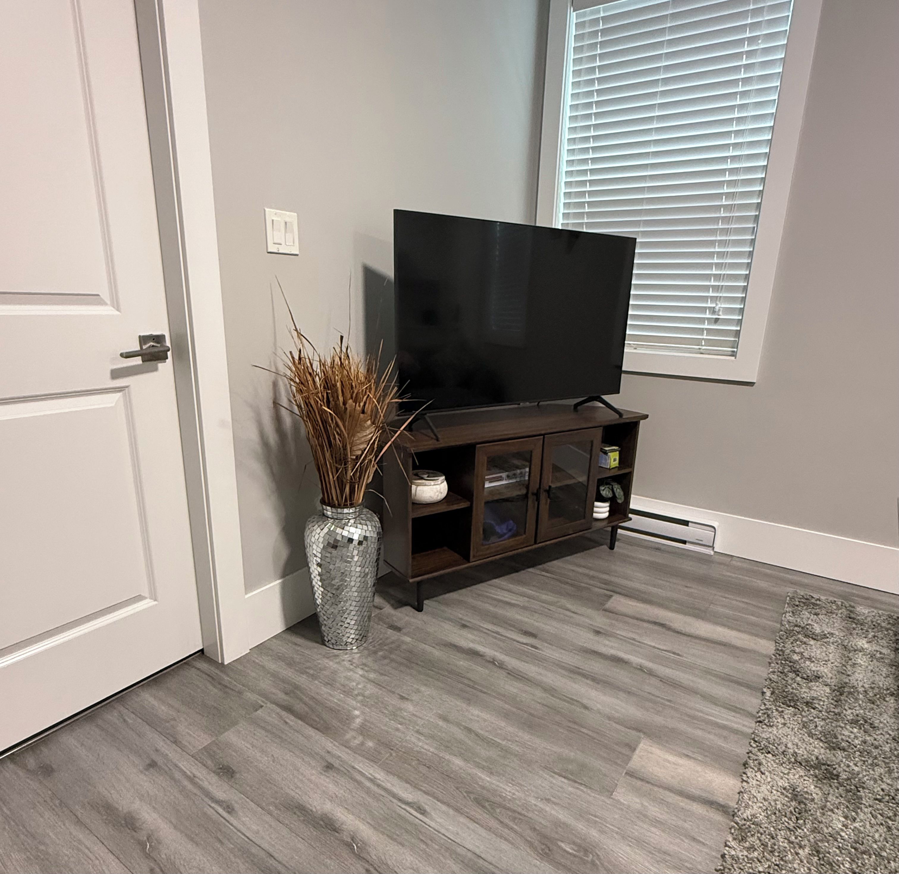A room with a television on a wooden stand, a decorative vase with dried plants, and a window with blinds.