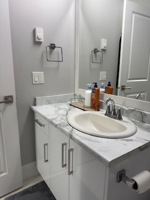 Een badkamer met een witte wastafel, een marmeren aanrechtblad en een spiegel erboven. Er staan toiletartikelen op het aanrechtblad en een handdoekhouder aan de muur.