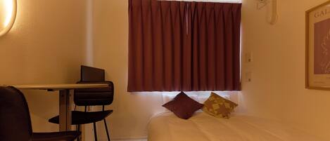 A hotel room with a bed, a desk, a chair, a window with curtains, and an air conditioning unit.