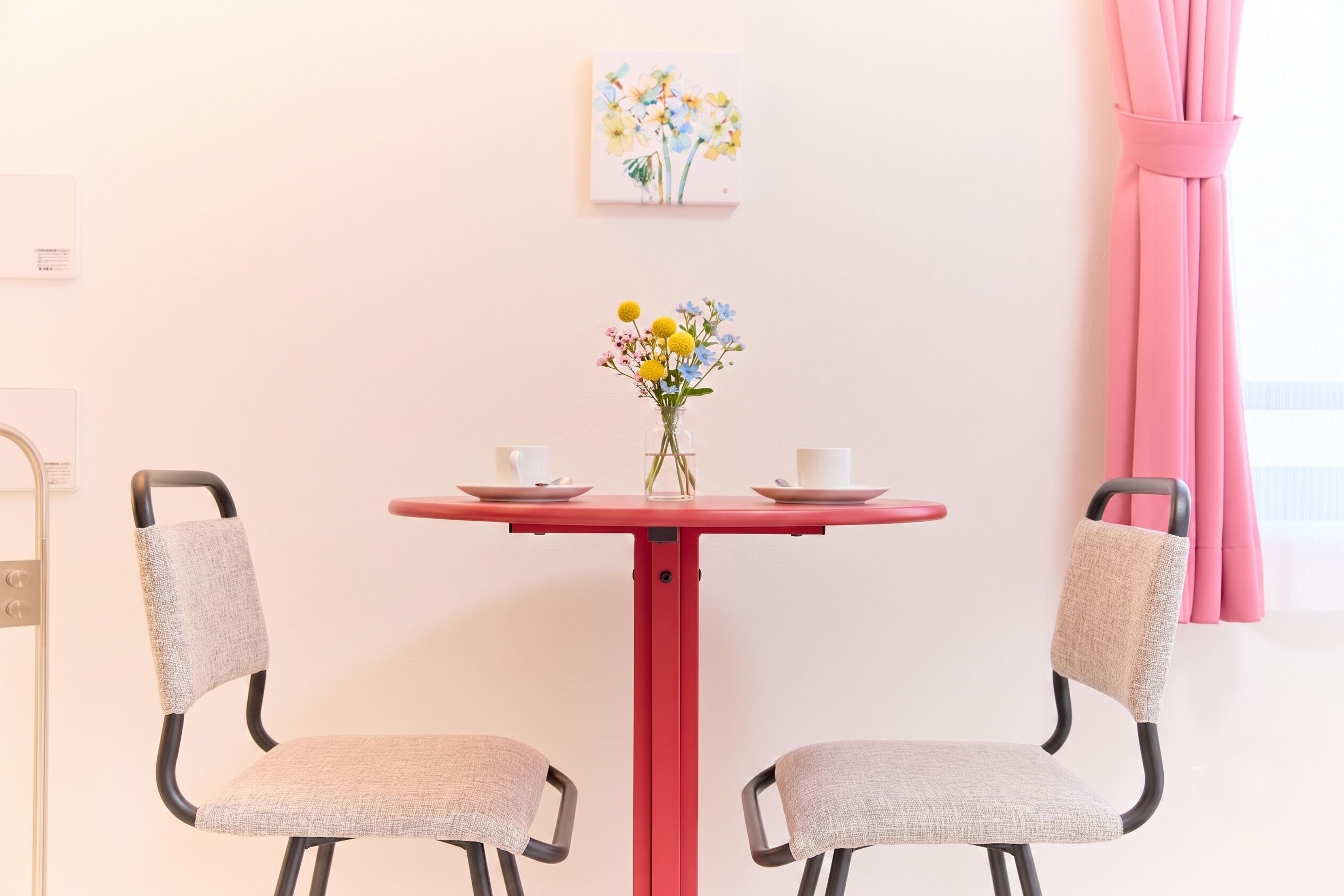 Um quarto pequeno com uma mesa vermelha, duas cadeiras, um vaso com flores e um quadro na parede.
