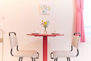Una stanza piccola con un tavolo rosso, due sedie, un vaso con fiori e un quadro appeso al muro.