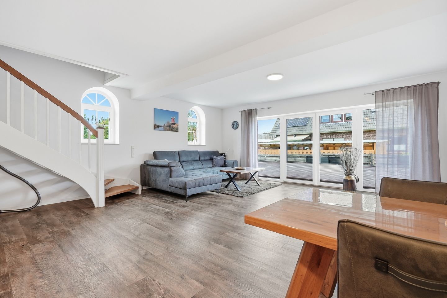 A spacious living area with a wooden staircase, a blue sofa, a dining table, and large windows with a view of buildings.