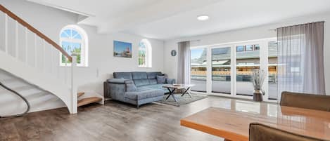 A spacious living area with a wooden staircase, a blue sofa, a dining table, and large windows with a view of buildings.