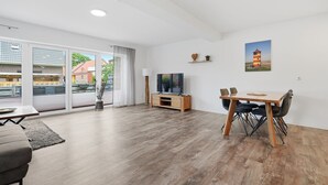 A modern living room with a flat-screen TV, a dining table with chairs, and a large window with a view of buildings and greenery. - To the Anchorage - To the Anchorage: Vacation near Greetsiel with a large terrace (Krummhörn)