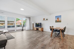 Ein modernes Wohnzimmer mit Flachbildfernseher, einem Esstisch mit Stühlen und einem großen Fenster mit Blick auf Gebäude und Grünflächen.