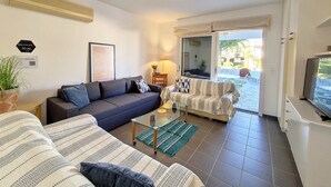 A living room with a sofa, a patterned couch, a coffee table, a TV, and a sliding glass door leading to an outdoor area. - Cosy Garden Flat, Beach, Pool – Mountain View, Private Pool, Air Conditioning (Akti Idras)