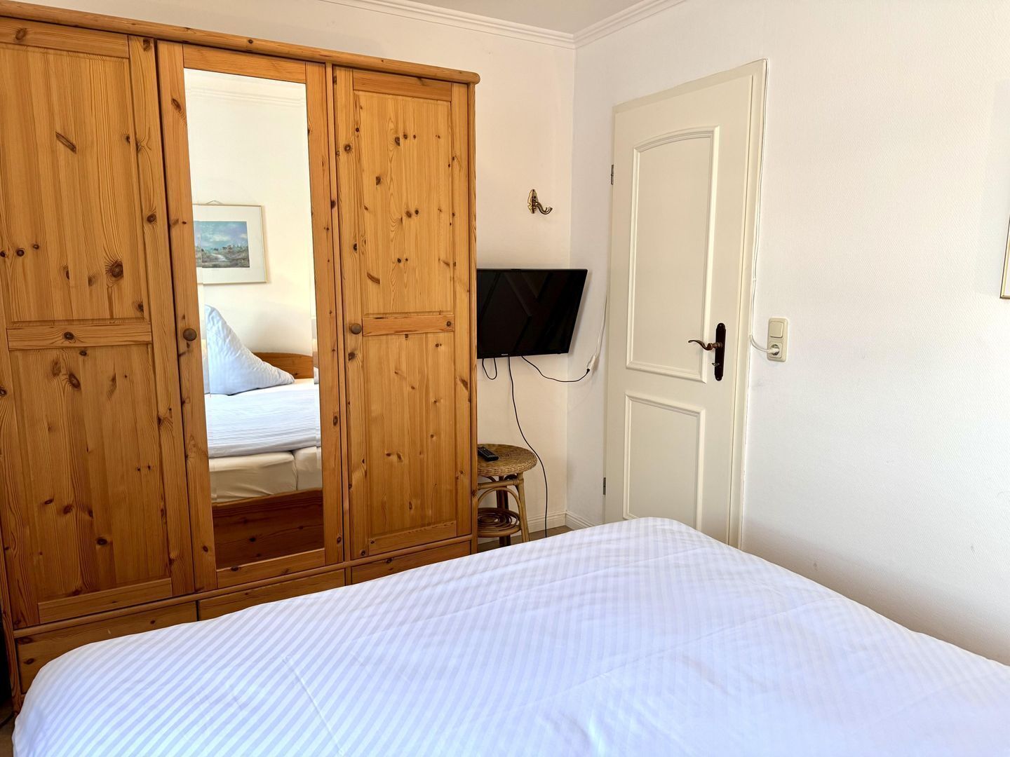 A bedroom with a bed, a wooden wardrobe, a television mounted on the wall, and a door with a handle.
