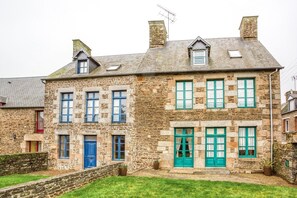 Un bâtiment en pierre doté de nombreuses fenêtres et portes, d’un petit jardin et d’une cheminée bien visible.
