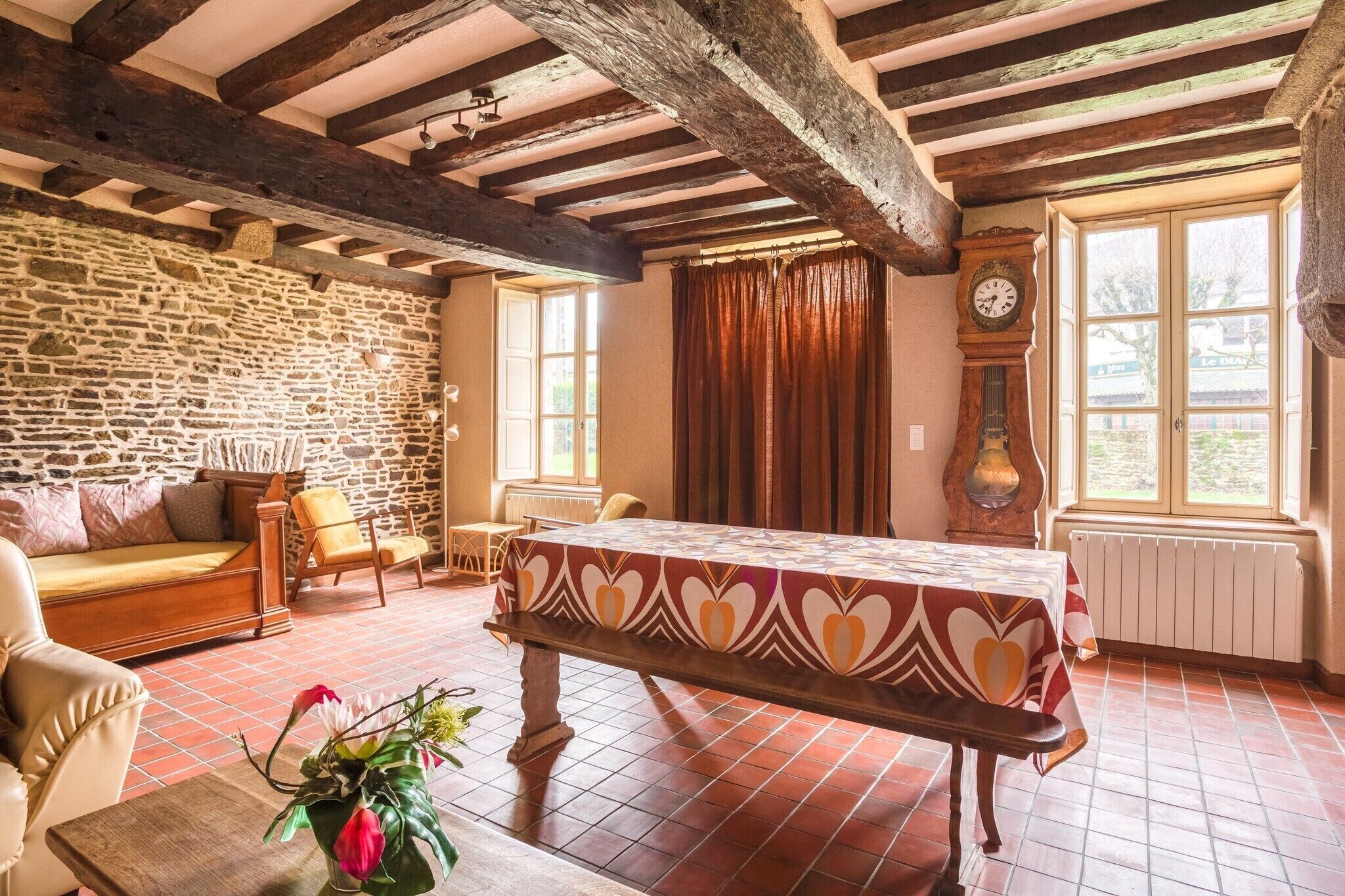 A traditional room with exposed wooden beams, a stone wall, a wooden table with a patterned tablecloth, a sofa, and a clock.