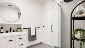 Ein modernes Badezimmer mit einem weißen Waschtisch, einer Duschkabine aus Glas und einem schwarzen Regal mit Pflanzen.
