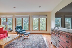 Ein Zimmer mit einem roten Sofa, einem blauen Sessel, einem Holztisch, einem gemusterten Teppich, einer Kommode mit mehreren Schubladen und großen Fenstern mit Jalousien.