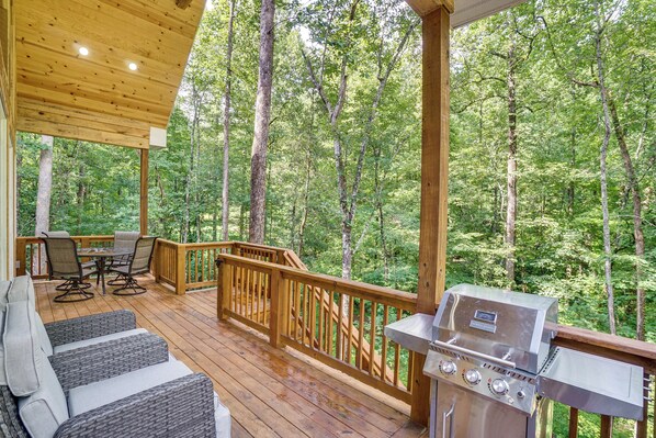 Une terrasse couverte, aménagée avec du mobilier d’extérieur, un barbecue et offrant une vue sur une forêt.