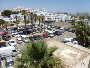 Een parkeerplaats met meerdere auto's en een paar bestelwagens, omgeven door palmbomen en gebouwen met witte gevels en blauwe koepels.