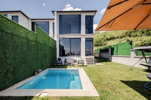 A modern two-story house with a swimming pool, a patio with chairs, and a large umbrella.