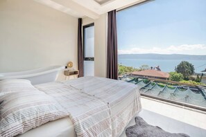 Ein Schlafzimmer mit großem Fenster, einem Bett und Blick auf ein Gewässer und die Landschaft.