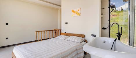 Une chambre Ă coucher avec un lit, une tĂŞte de lit en bois, une baignoire blanche et une fenĂŞtre donnant sur un paysage verdoyant.