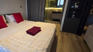 Habitación de hotel con cama, toalla roja, mesita de noche, armario y baño visible a través de una puerta de cristal.