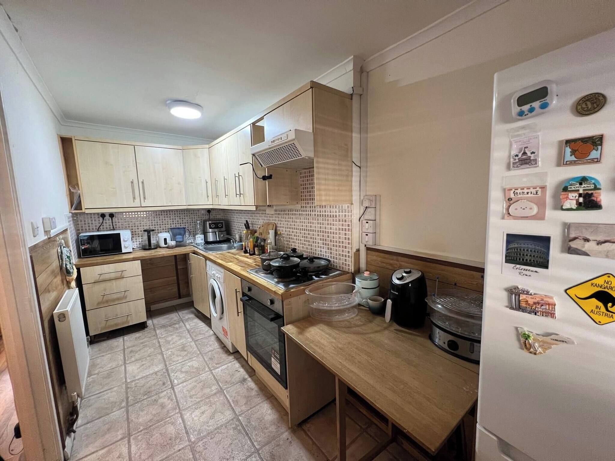 A compact kitchen with wooden cabinets, a microwave, a washing machine, and a refrigerator adorned with magnets.