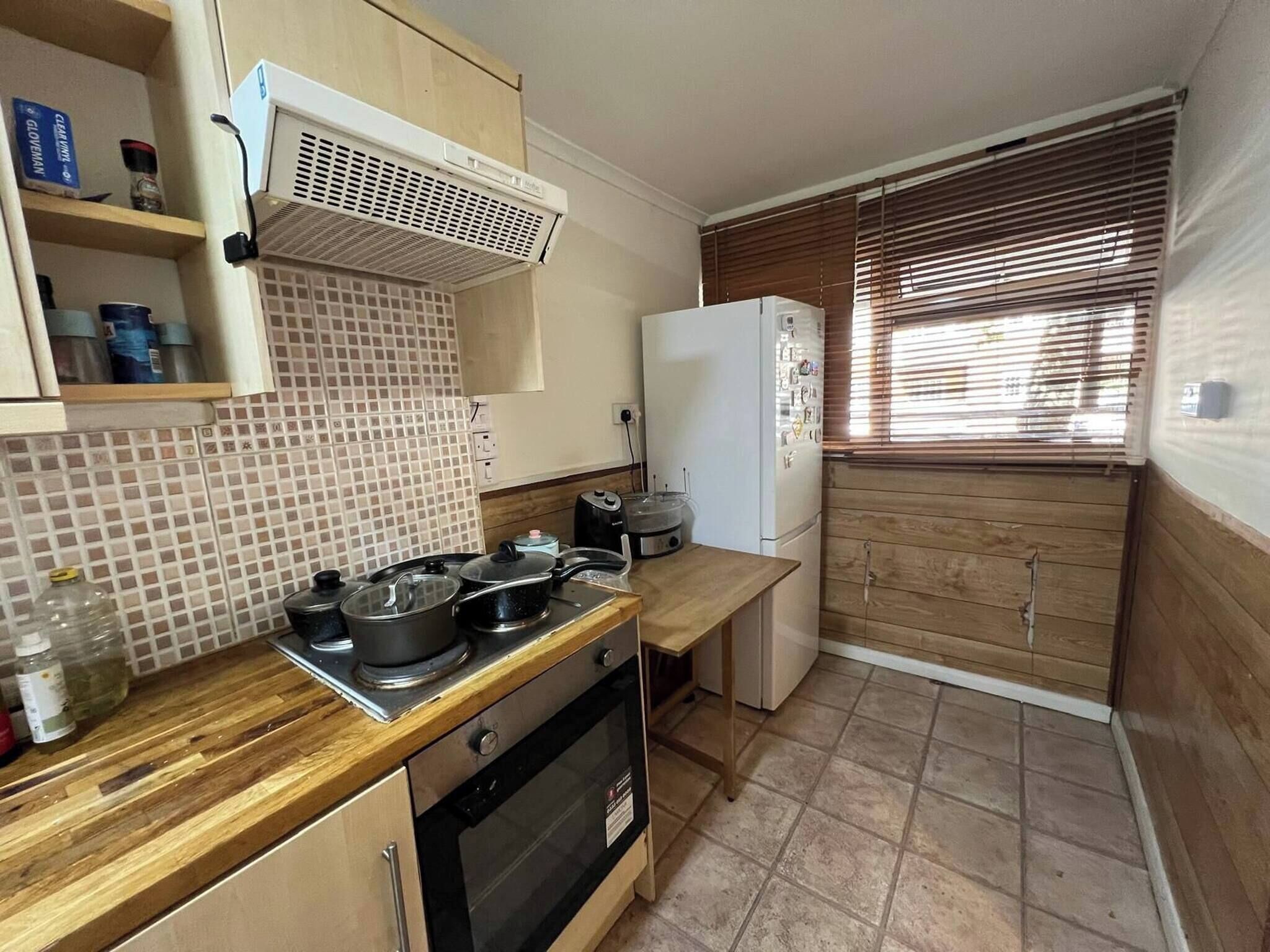 A compact kitchen with a stove, oven, and a refrigerator. There is a window with blinds and a tiled backsplash.